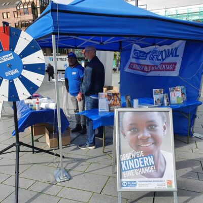 A blue tented booth with a spinning prize wheel, pamphlets, and posters promoting child sponsorship. A large sign in front features a smiling child and reads, Geben Sie Kindern eine Chance!.